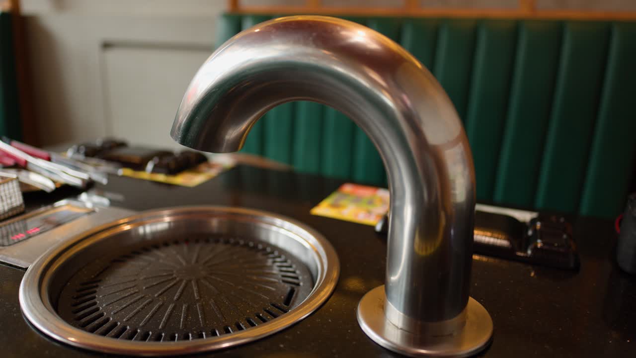 Stainless steel exhaust vent rotates above empty Korean grill pan in warmly lit restaurant booth
