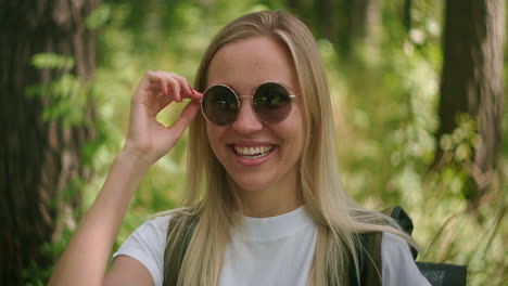 a young female traveler in sunglasses looks directly at the camera and smiles flirting a traveler with a backpack in the park and in the forest in slow motion