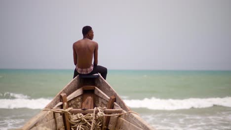 rear view of unidentifiable african male sitting at the edge of a canoe relaxing and facing the ocean and opening his hands