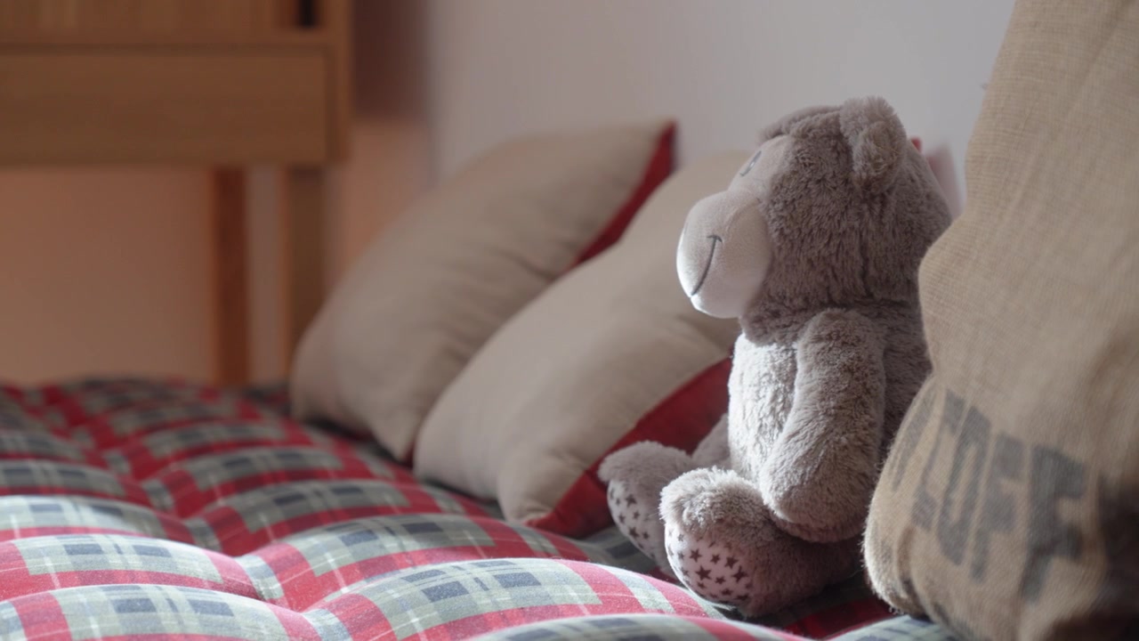 Orsetto di peluche che riposa su un letto rosso con cuscini in un accogliente interno di camera da letto sotto una morbida luce naturale