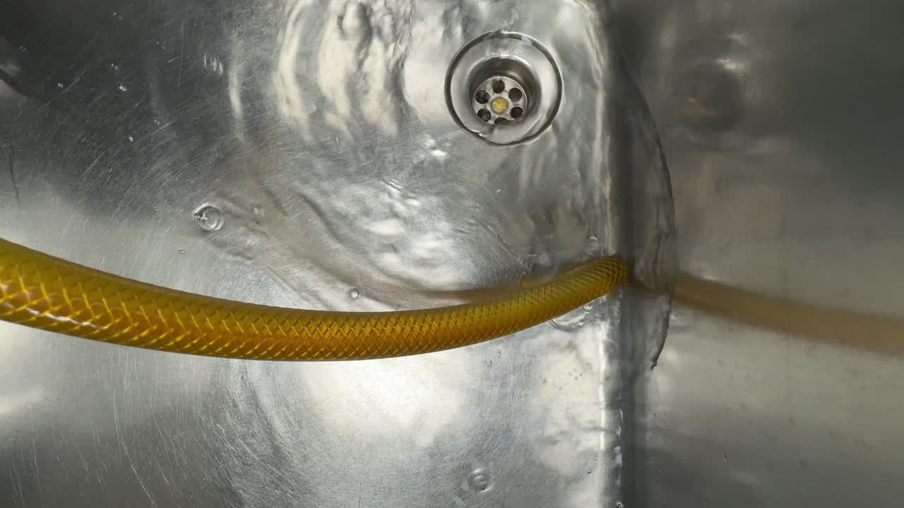 A close-up view of a stainless steel sink with water flowing around a yellow flexible hose. The metallic surface reflects light, creating abstract patterns and textures