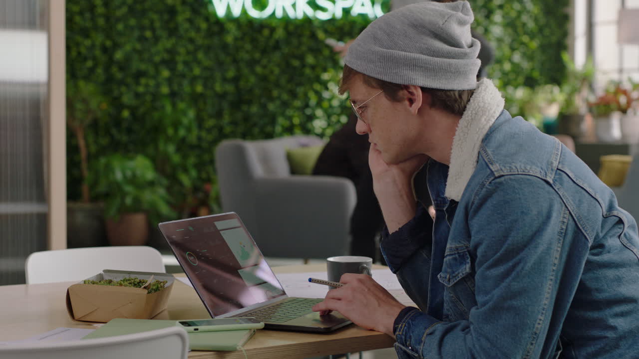 молодой кавказский студент использует ноутбук, работает над творческим проектом, просматривает онлайн-исследования, наслаждается учебой в современном офисном рабочем пространстве