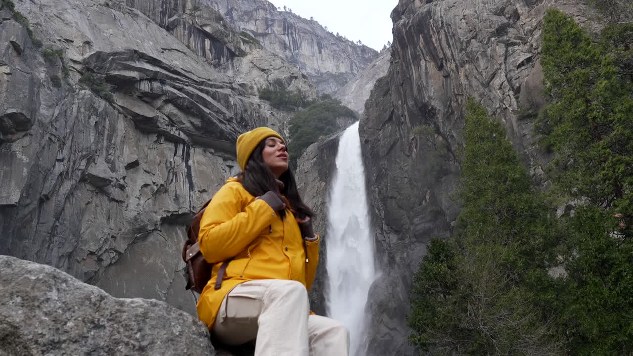 Happy Woman Sitting by a Majestic Waterfall in a Mountainous Landscape