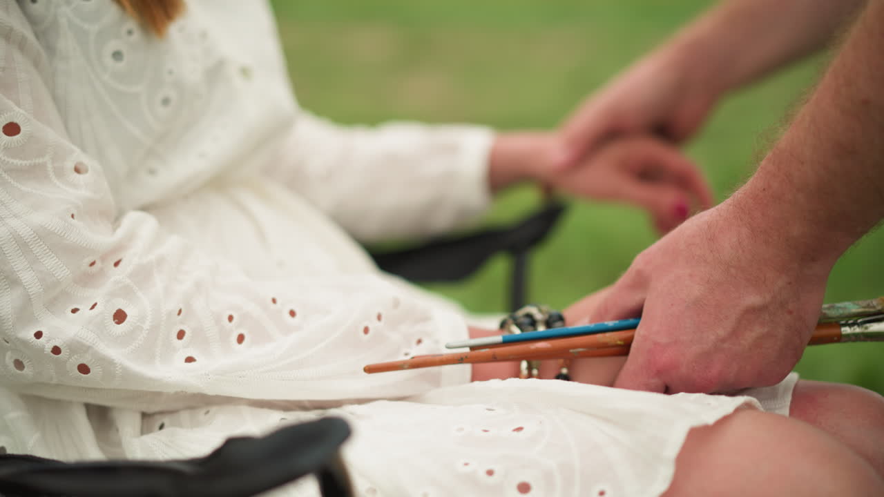 평화로운 야외 장면에서, 체크 셔츠를 입은 예술가는  드레스를 입은 앉아있는 여성의 손을 조심스럽게 조정하면서 질을 부드럽게 들고 있습니다.