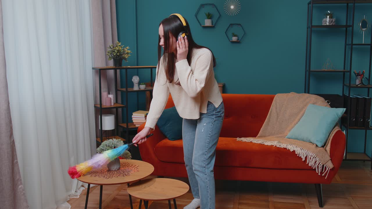 Young dancing woman dusting furniture alone caring for hygiene using colorful duster in room at home