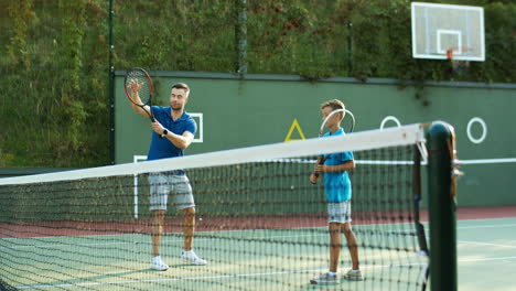 Man-Teaching-His-Teen-Son-How-To-Play-Tennis-And-Then-Giving-High-Five-On-An-Outdoor-Court-In-Summer