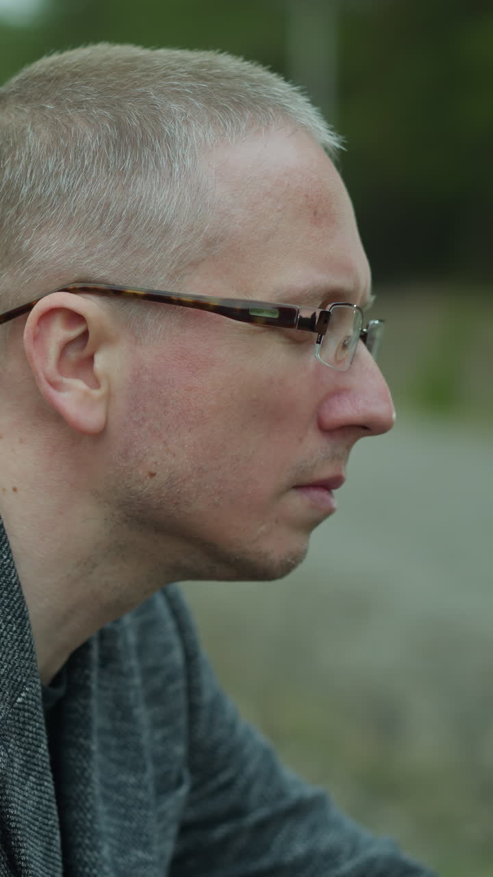 una vista cercana de un hombre contemplativo con gafas, mirando hacia otro lado mientras está sentado junto a una vía ferroviaria, el fondo sereno del bosque contrasta con su expresión pensativa