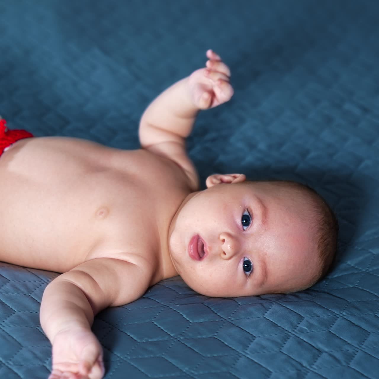 Blue-eyed Caucasian baby boy wearing red shorts and socks only tosses little feet. Beautiful child lying on the grey background
