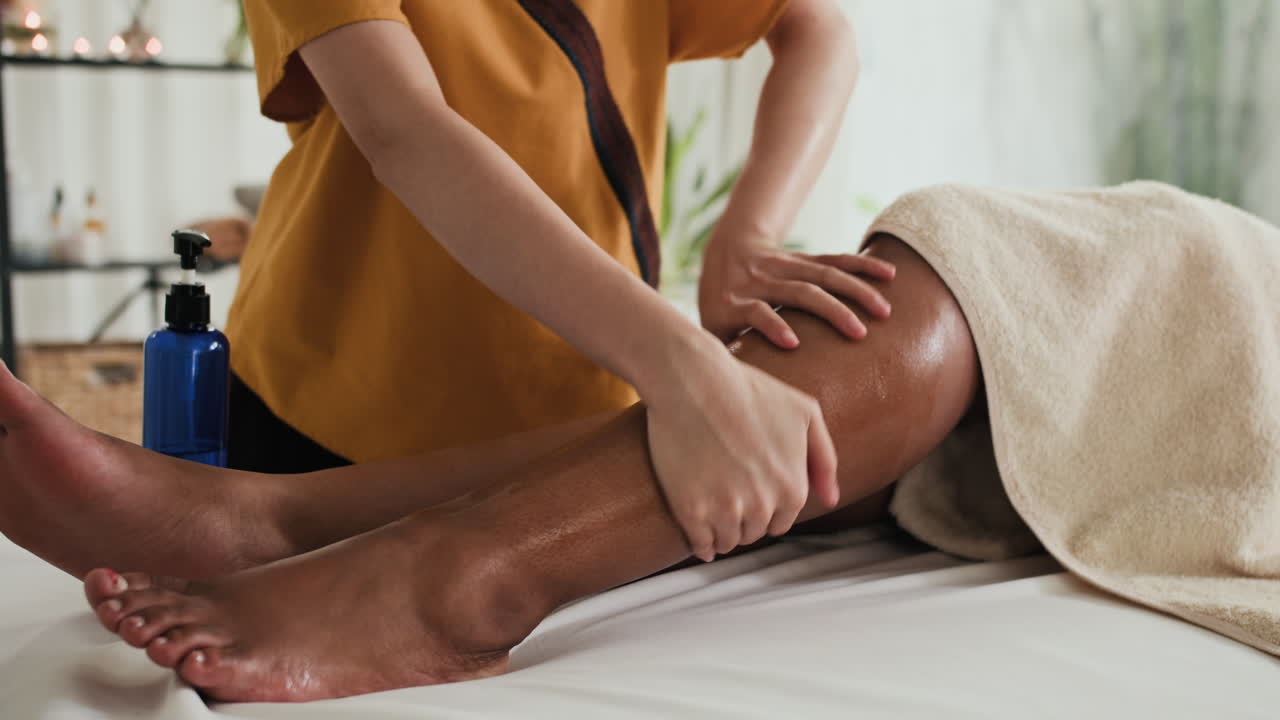 Hands of Masseuse Hydrating Skin on Legs of Client in Beauty Studio
