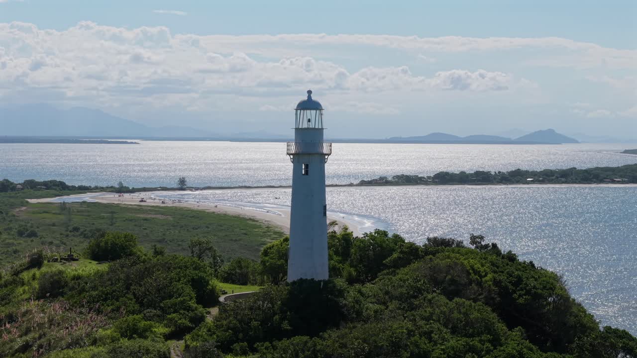 Valkoinen Faro das Conchas -majakka (Farol das Conchas) seisoo Ilha do Melillä lähellä Paranaguáa, Paranáa, Brasiliassa, Atlantin rannikon pensaikkojen ympäröimänä, taustalla ranta- ja lahdenvedet, lennokin kiertokuva.
