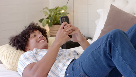 young man relaxing on bed, smiling while using smartphone at home