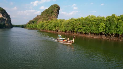 Aerial-drone-of-a-caucasian-male-tourist-standing-on-a-Thai-longtail-boat-motoring-down-river-with-large-green-limestone-mountain-rocks-surrounded-by-mangrove-forests-during-sunset