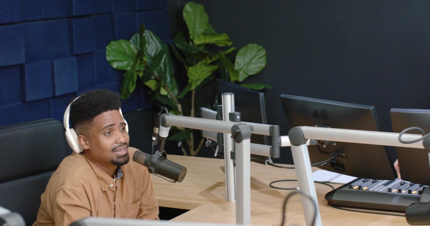 Co-hosts wearing headsets, woman in denim jacket speaking, camera panning to man at mic, recording