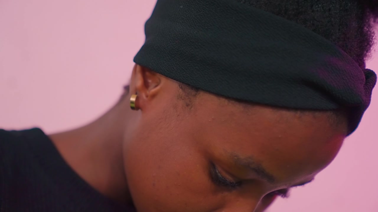 Primo piano di una donna di colore che guarda in basso pensierosa in una luce rosa da studio, indossa un berretto e un piccolo orecchino con una leggera inclinazione della testa e uno sguardo verso il basso, un'atmosfera intima e contemplativa, ombre morbide accentuano la pelle.