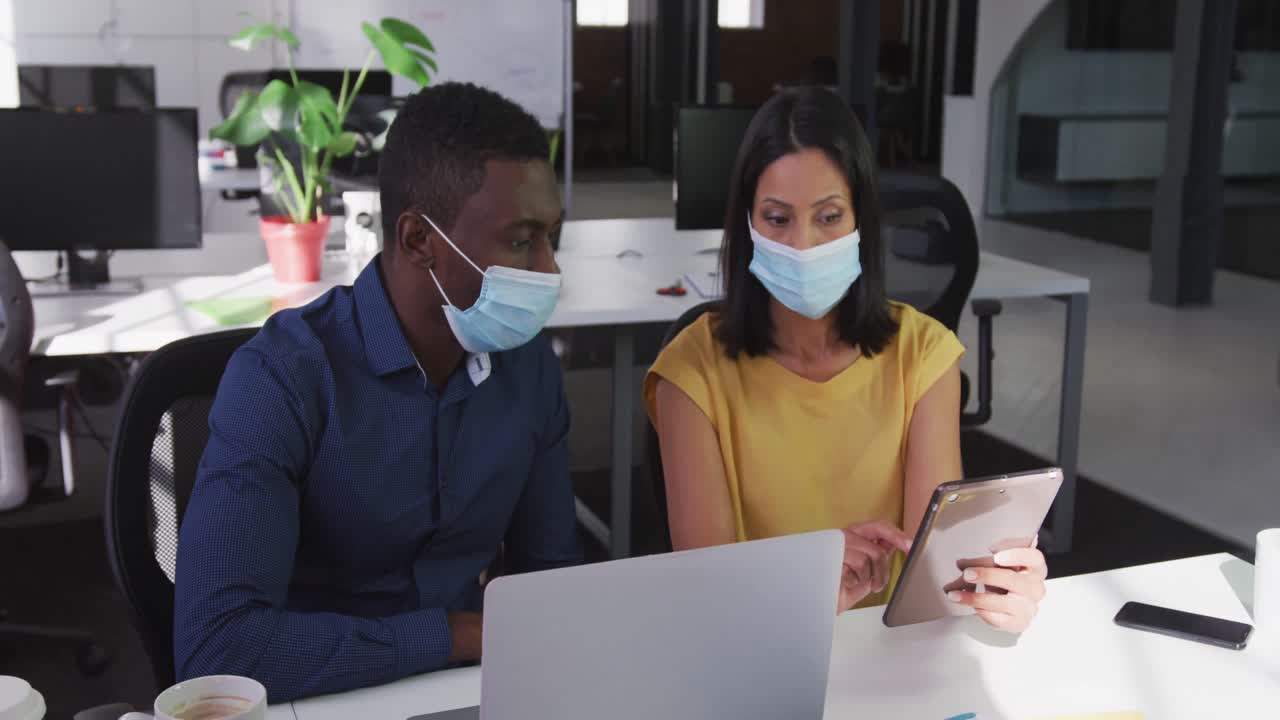 Diverse male and female business colleagues wearing face masks sitting at desk using tablet