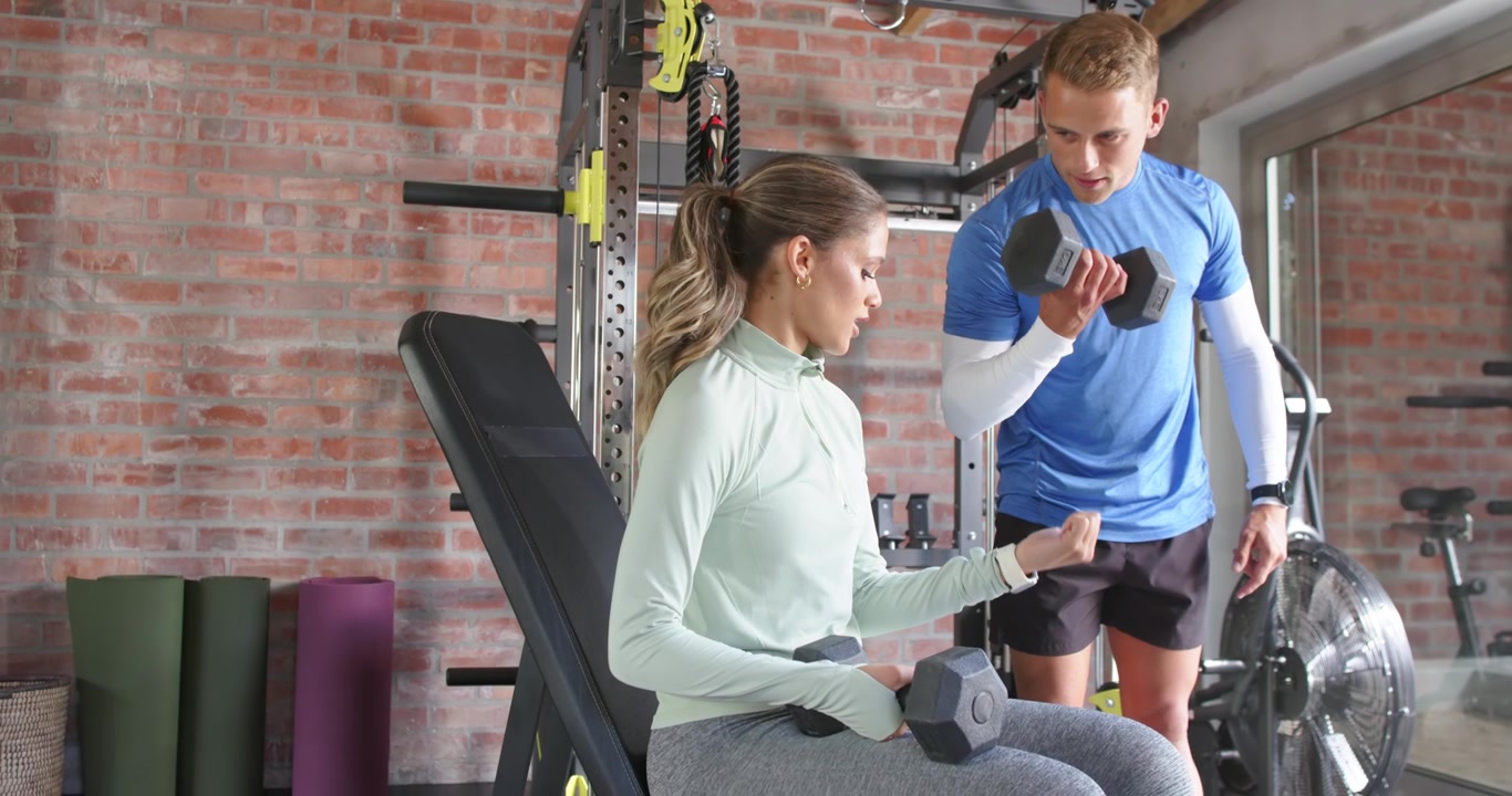 Male trainer approaching guiding woman client pointing at wrist showing curl with weights for form
