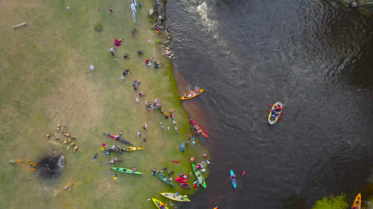 Top down aerial view of SUP mega paddleboards and colorful kayaks going through foamy river rapids on a narrow river from underneath a bridge during Võhandu marathond while spectators watch on sides.