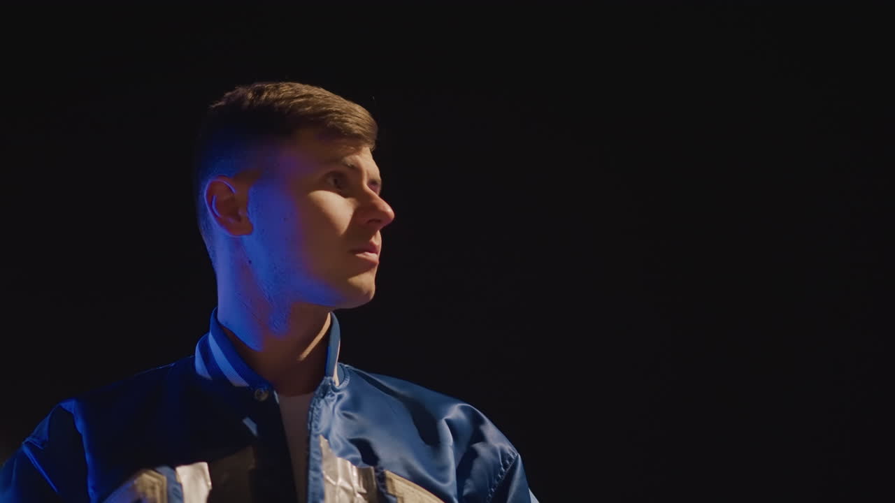 네온 불빛 아래 차 안에 있는 젊은 운전자, 바시티 재킷과 보라색 실내 조명, 창문에 기대다가 텅 빈 주차장으로 나와 생각에 잠긴 표정으로 위를 올려다보는 영화 같은 로우 키 조명