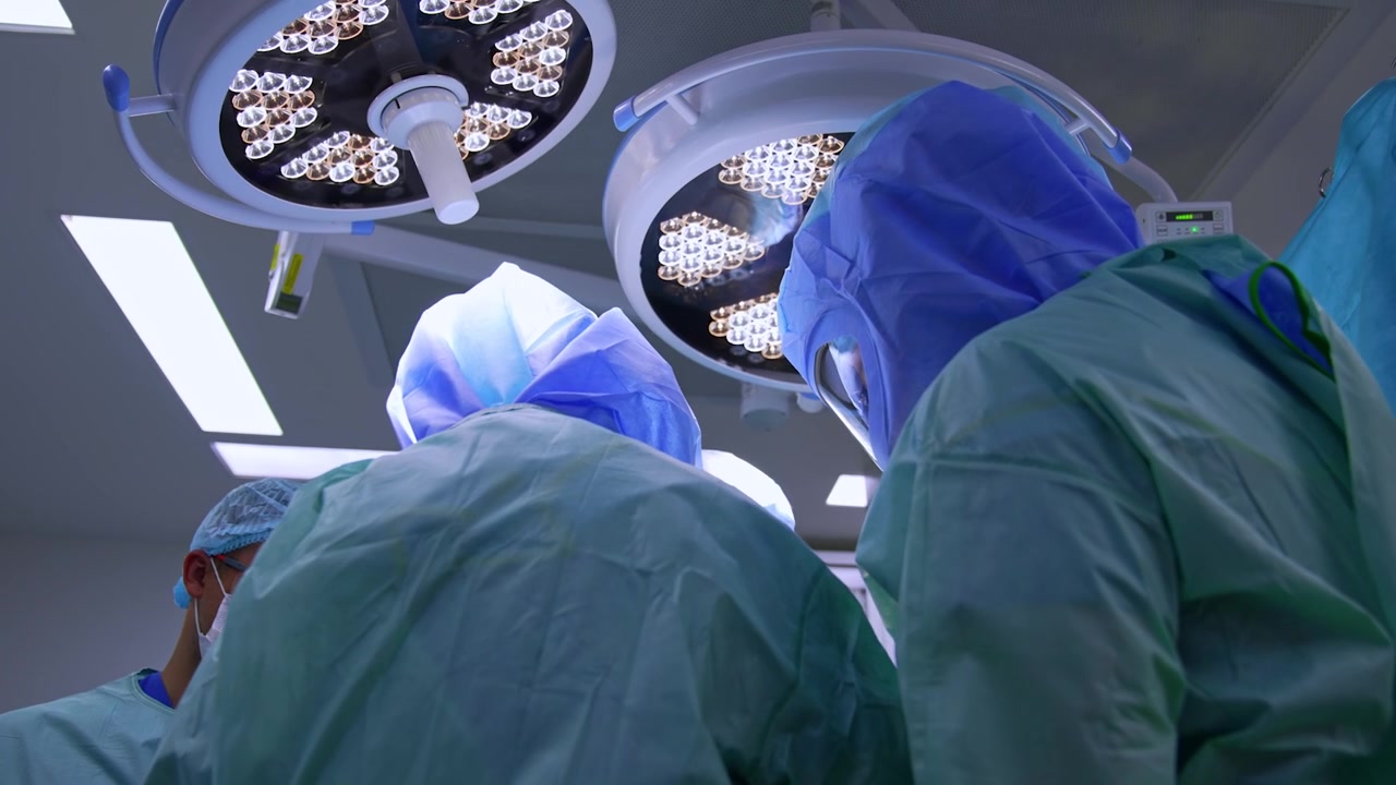 Backs of two surgeons in blue robes and helmets performing surgery. Low angle view on the medics under the round illumination equipment.