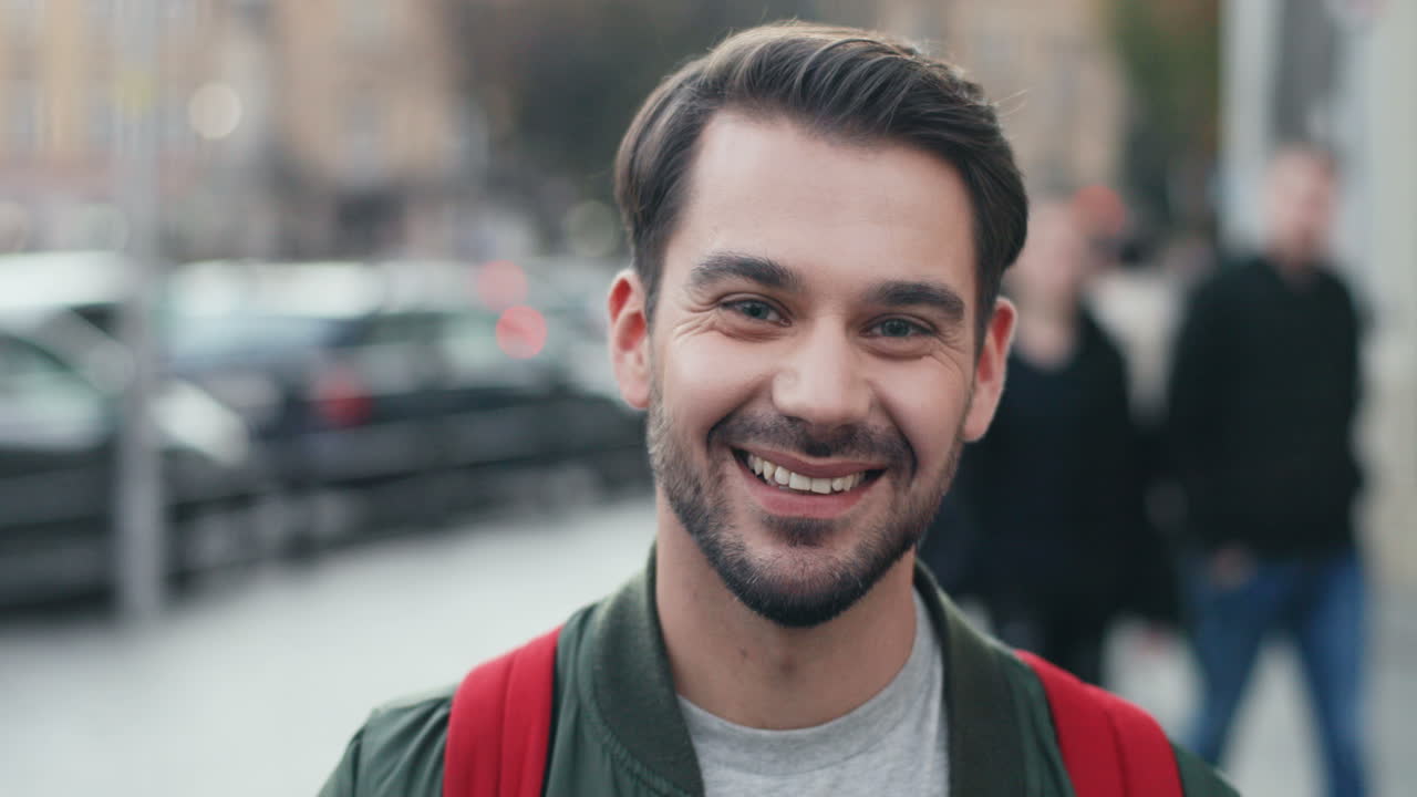 Portrait of young Caucasian man with backpack looking aside in the street, then turns his face and smiles at the camera