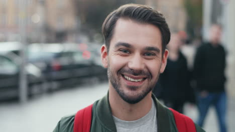 Portrait-of-young-Caucasian-man-with-backpack-looking-aside-in-the-street,-then-turns-his-face-and-smiles-at-the-camera