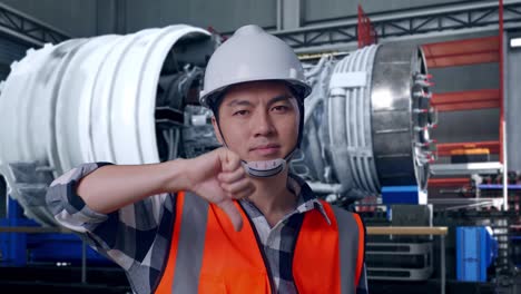 close up of asian male engineer with safety helmet showing thumbs down gesture and shaking his head while standing with airplane engine maintenance conducted, analytics checking the turbine