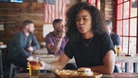 young woman watching game on screen in sports bar eating burger and fries and drinking beer