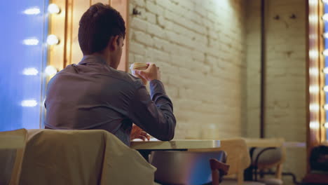 Student in formal shirt drinks coffee while sitting alone inside cozy cafe near glowing window, with jacket on chair, phone on table, and backpacks on bench, surrounded by brick walls and soft evening light