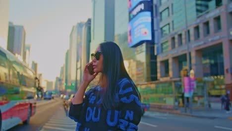 Hermosa-Modelo-De-Mujer-Asiática-Oriental-En-Una-Gran-Ciudad-Urbana-Con-Gafas-De-Sol-Y-Llamadas-De-Teléfono-Celular-Y-De-Pie-Cerca-De-Rascacielos