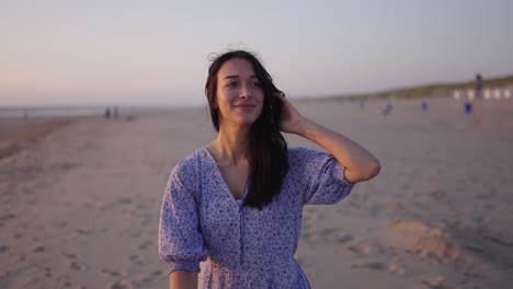 Attractive-smiling-brunette-in-summer-dress-holds-back-hair-walking-on-beach