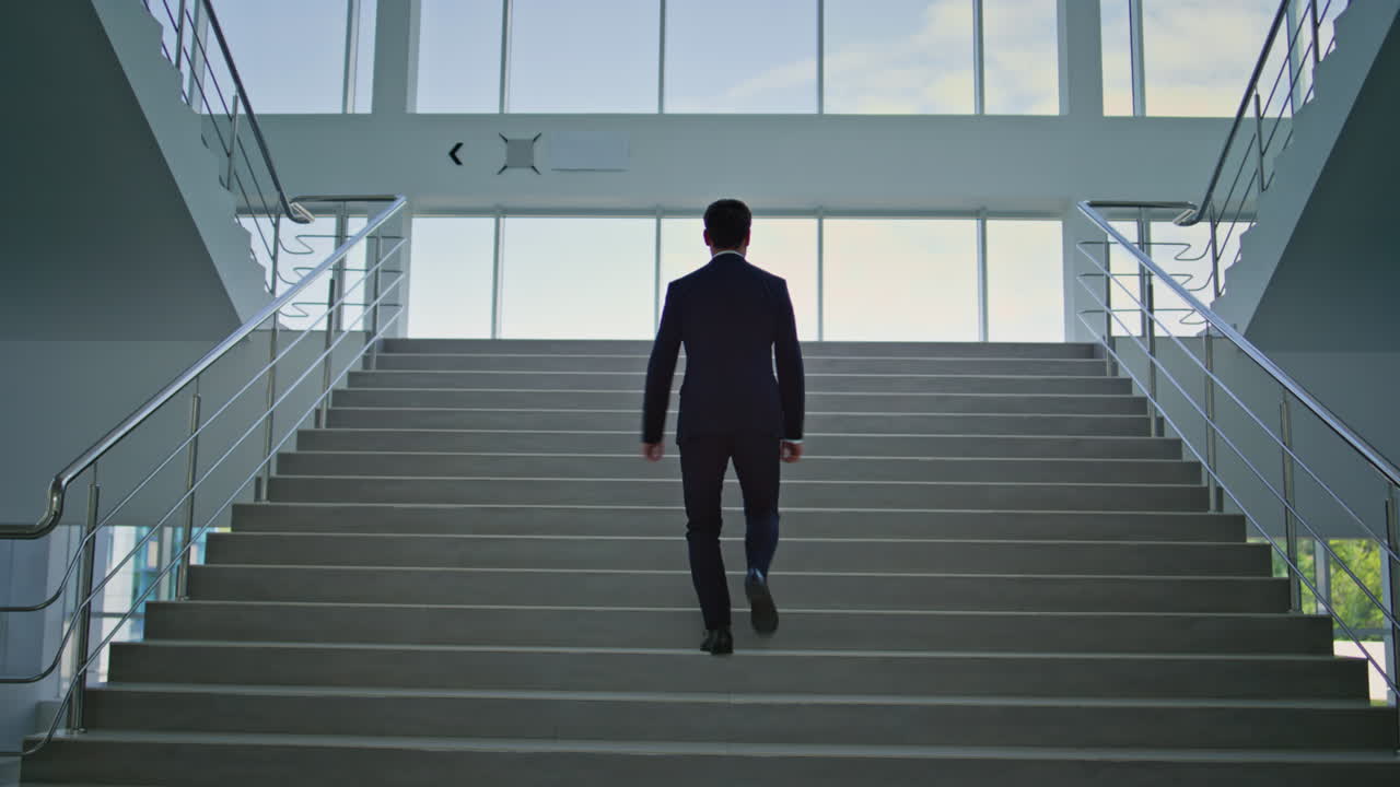 Back view businessman climbing modern glass stairway in bright office interior