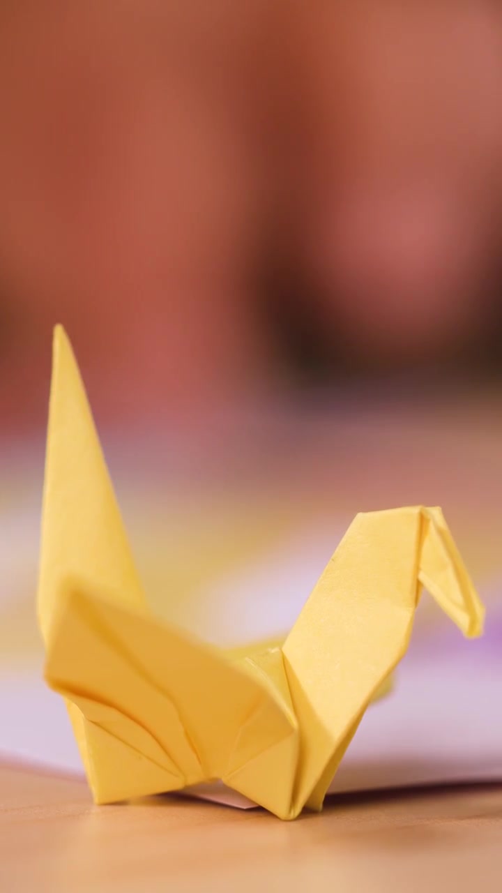 A finished yellow origami crane sits on a wooden table with blurred hands working behind