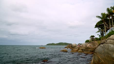 Pan-right-of-sea-waves-hitting-a-rocky-shore-near-a-resort-in-Bombas-and-Bombinhas-beaches,-Brazil