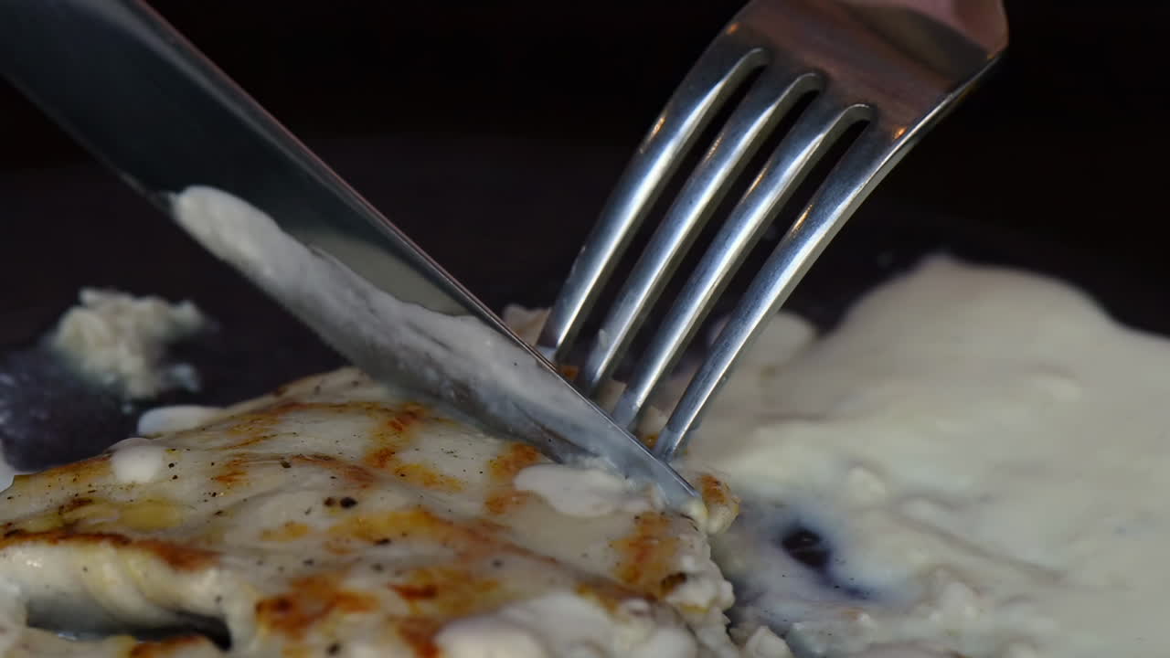 Nærbillede af en kniv og gaffel, der skærer et stykke grillet kylling med hvid sovs