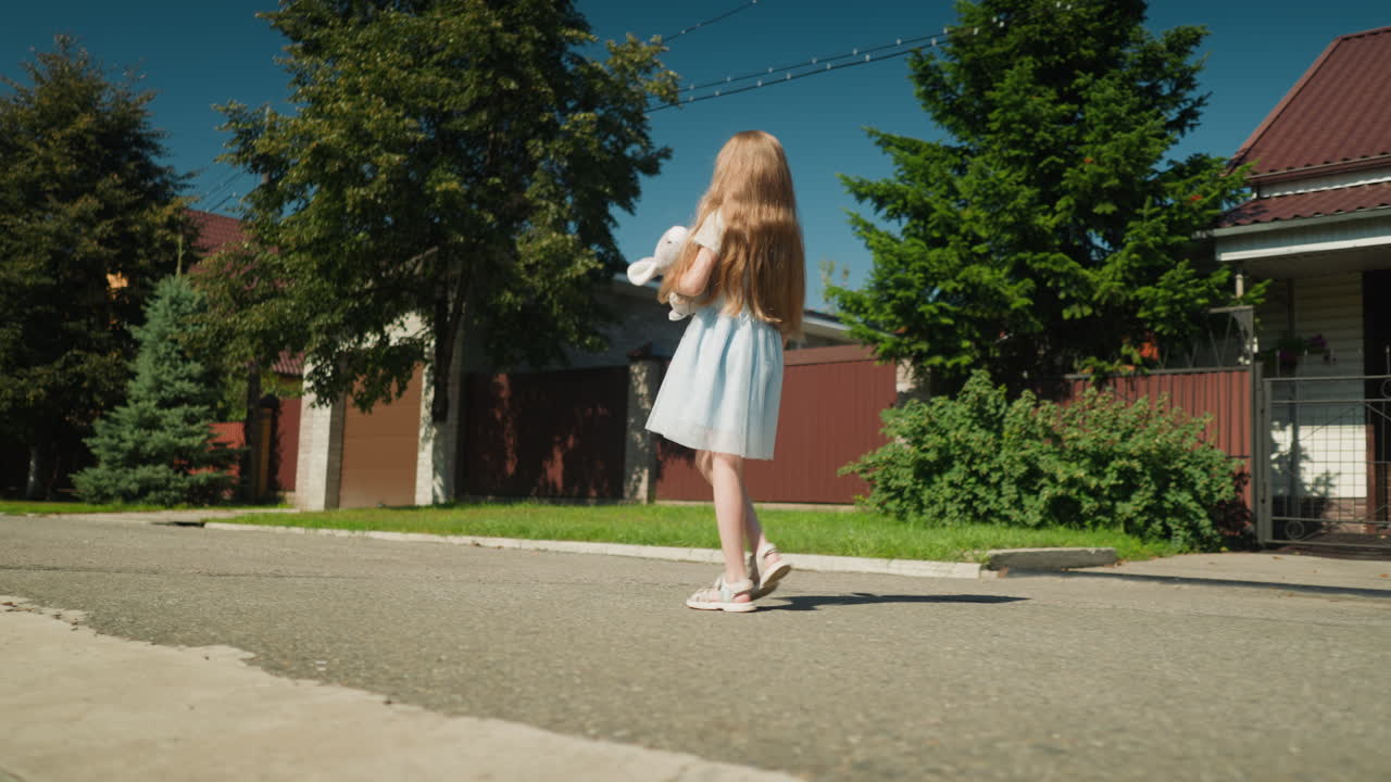 Side view of little girl with long hair wearing light dress holding stuffed rabbit toy while walking alone along quiet sunny street with tree shadows and residential houses in background