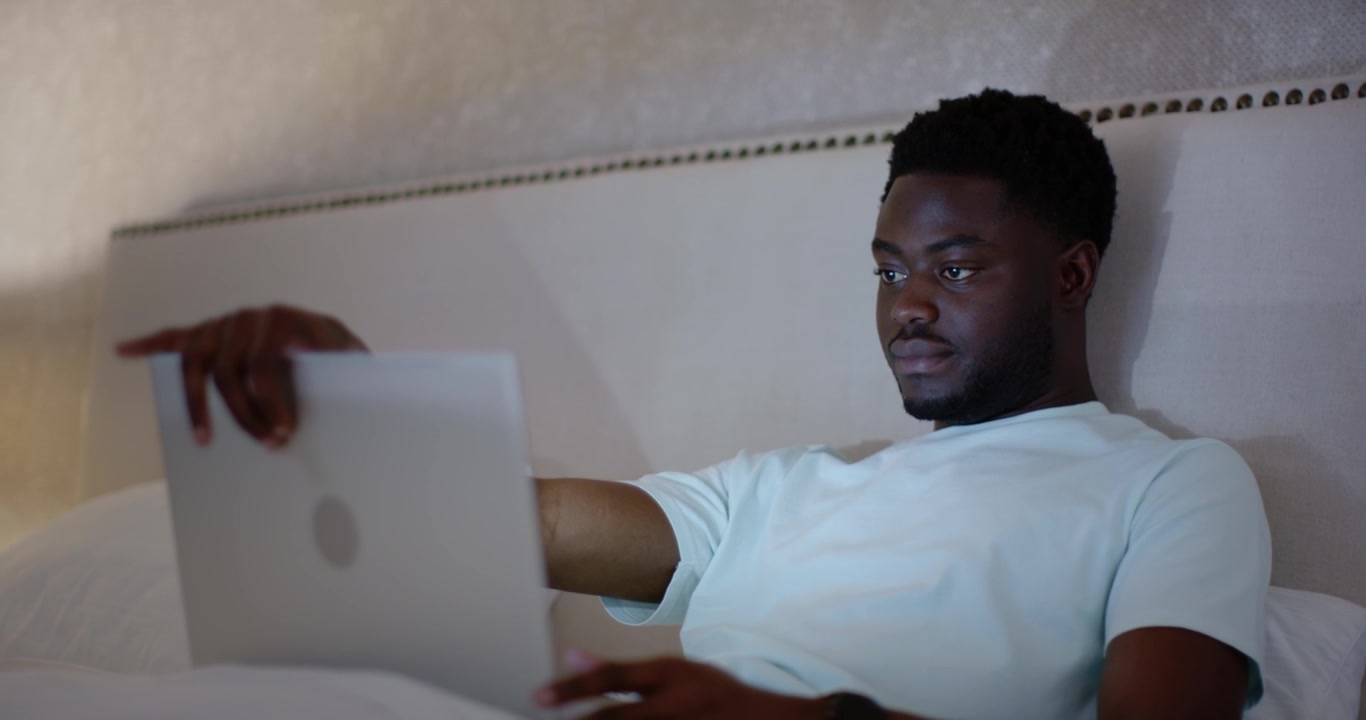 Afroamerikansk mand holder bærbar computer, åbner den i sengen ved sengegavlen, taster med ur på