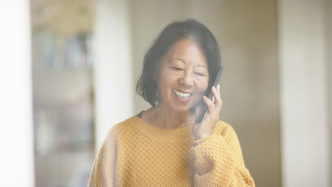 senior asian woman in yellow sweater smiling while talking on smartphone at home
