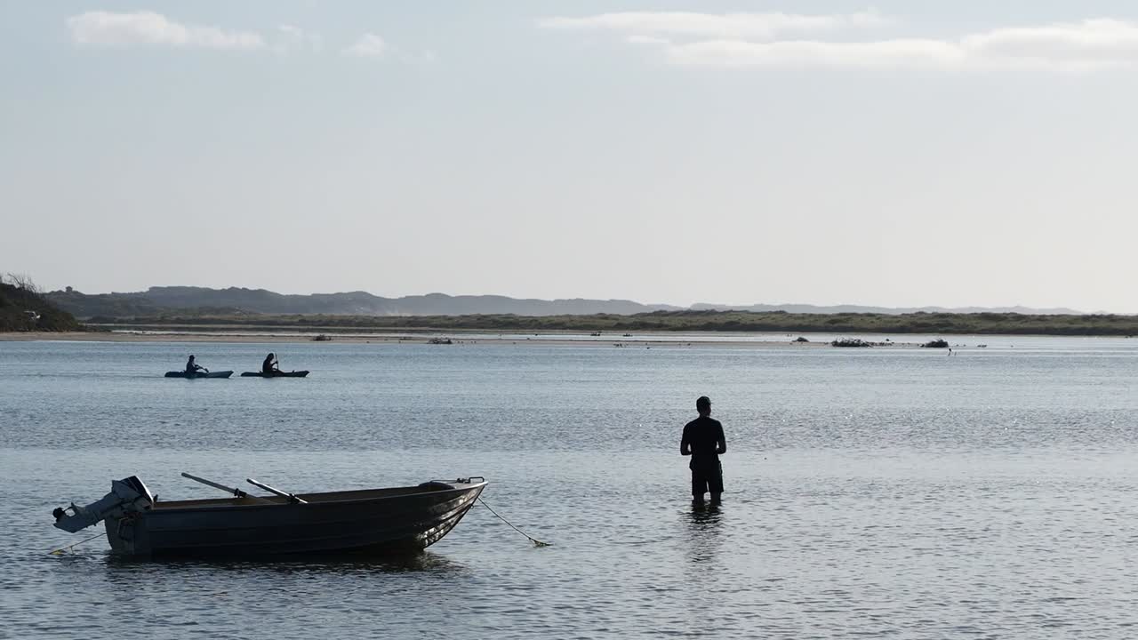 Silueta de pescador vadeando con bote auxiliar amarrado y kayakistas