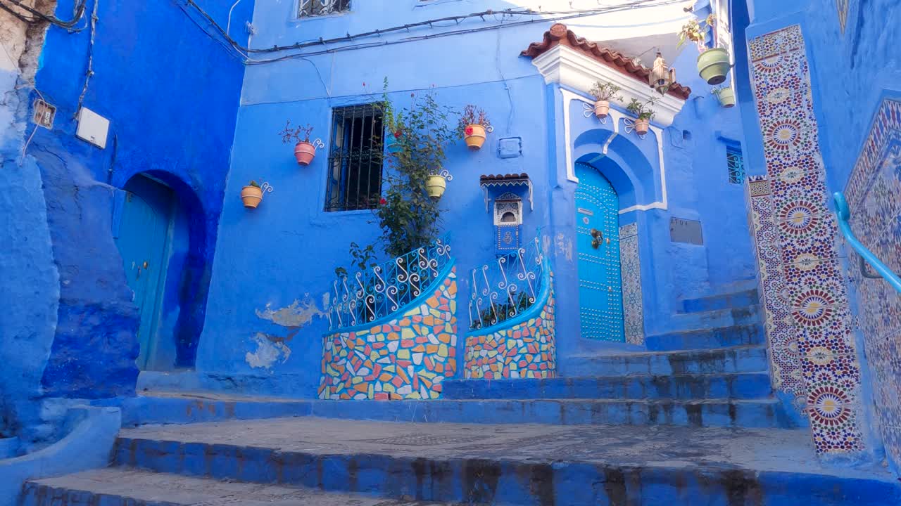 Tilt Down View Across Beautiful Blue Door With Two Small Flower Gardens Beside Narrow Street With Steps In Chefchaouen In Morocco