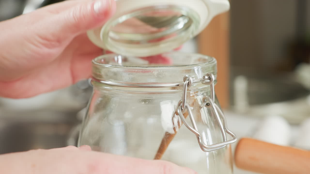 Close up of hands opening glass flour container with metal clip lid in kitchen, preparing to scoop flour using wooden spoon inside, surrounded by soft natural lighting and kitchen items
