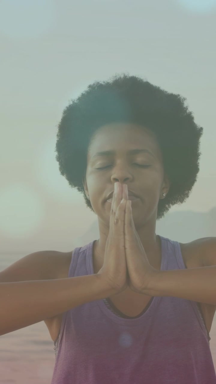 Vertical video: Mid adult woman exhaling and lowering palms to heart at shoreline, health practice