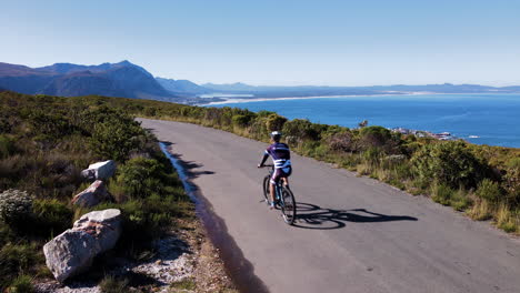 MTB-ride-on-mountain-road-through-fynbos-with-panoramic-ocean-views,-aerial