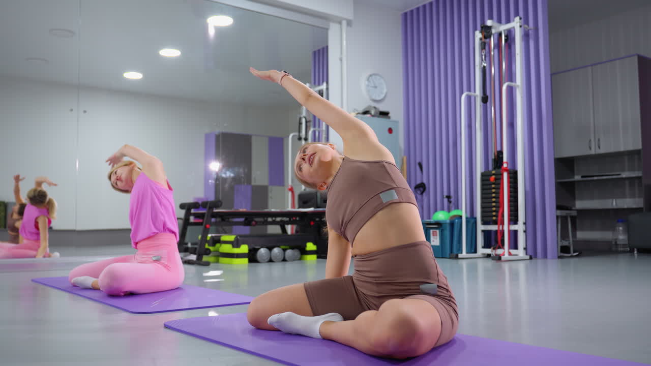 mujeres sentadas en esteras púrpuras realizando ejercicios de estiramiento de costillas, el fondo presenta varios equipos de fitness, incluidas bandas de resistencia y pesas, con un gran espejo que refleja sus movimientos