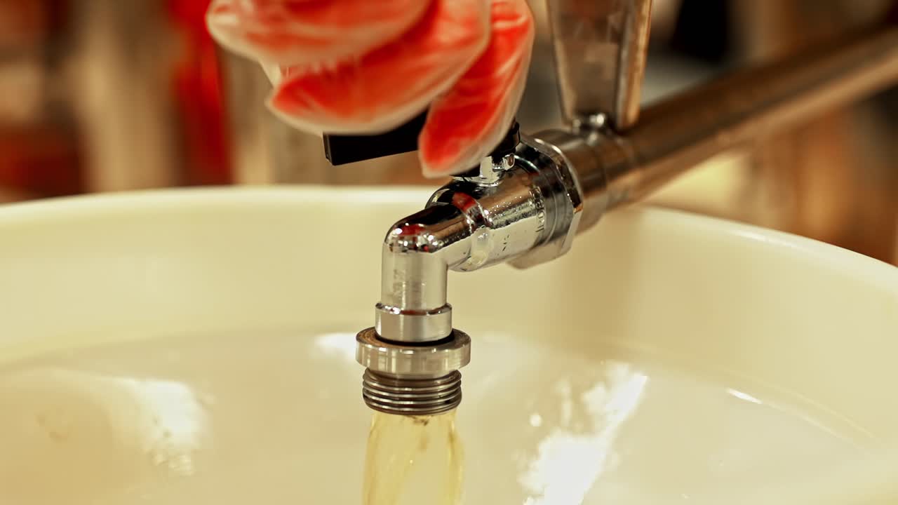 Running amber liquid flows from faucet into sink with clean bright surface and steady controlled stream, gloved hand adjusts speed