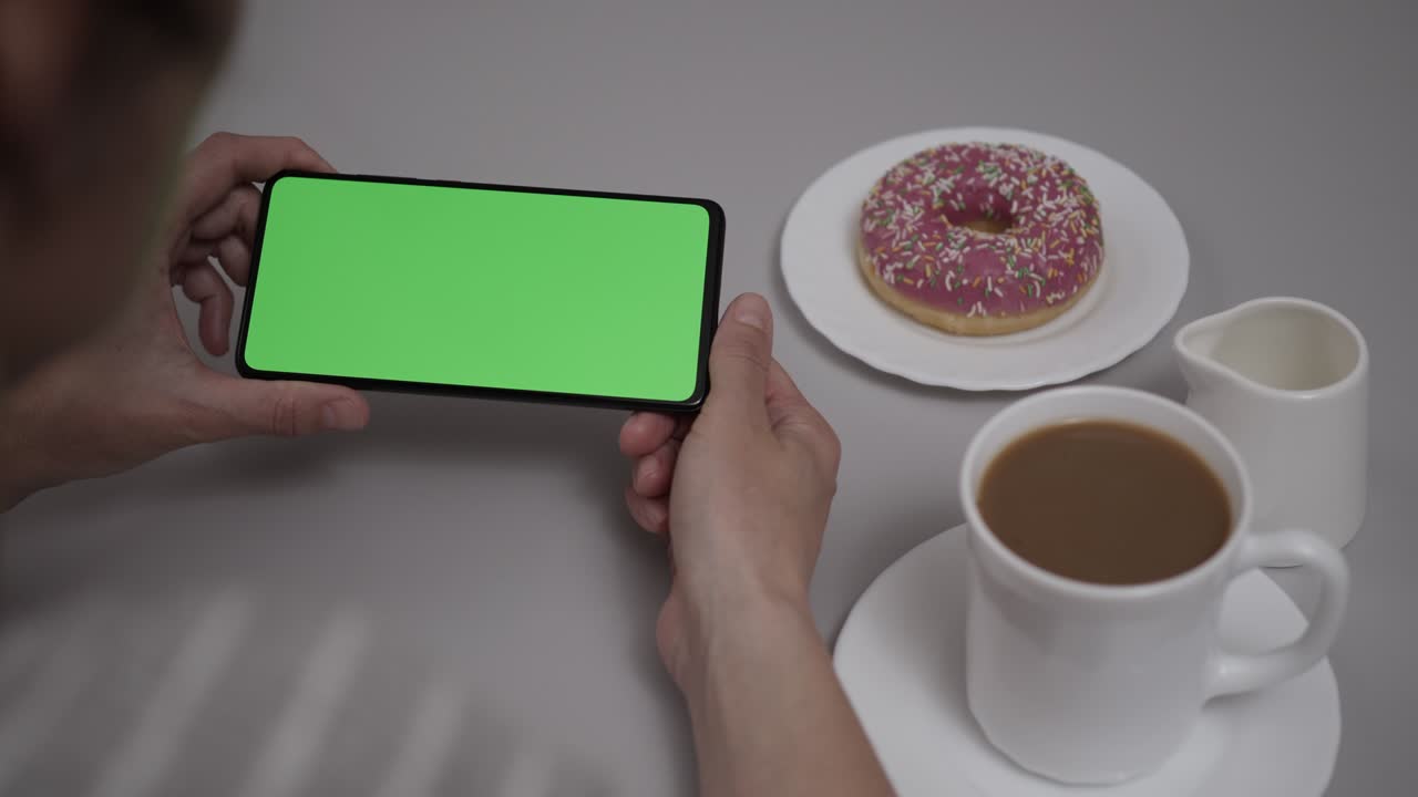mujer sentada en la mesa de café donut usando un teléfono inteligente con pantalla verde de clave de croma, desplazándose a través de las redes sociales medios de comunicación en línea tienda internet. teléfono inteligente en modo horizontal con maqueta de pantalla verde.