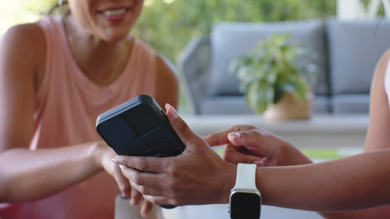 Diverse women sharing on patio holder with white watch angling phone as friend coming with bottle