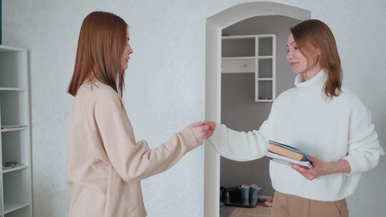 Due donne si salutano sulla soglia dell'appartamento scambiandosi quaderni e sorrisi, inquadratura media mostra un saluto informale con il pugno e una stretta di mano cortese mentre una tiene una pila di libri, scaffalature pulite e illuminazione soffusa creano un'atmosfera calda