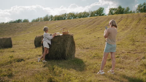la señora toma fotos de su elegante amiga de pie al lado de la bala de heno con la configuración de picnic en el campo pintoresco, la luz del sol destaca la atmósfera rural relajada