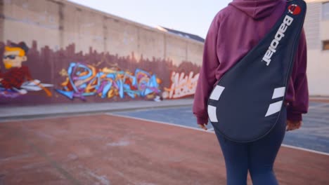 female tennis player entering an urban concrete tennis court carrying a tennis racket