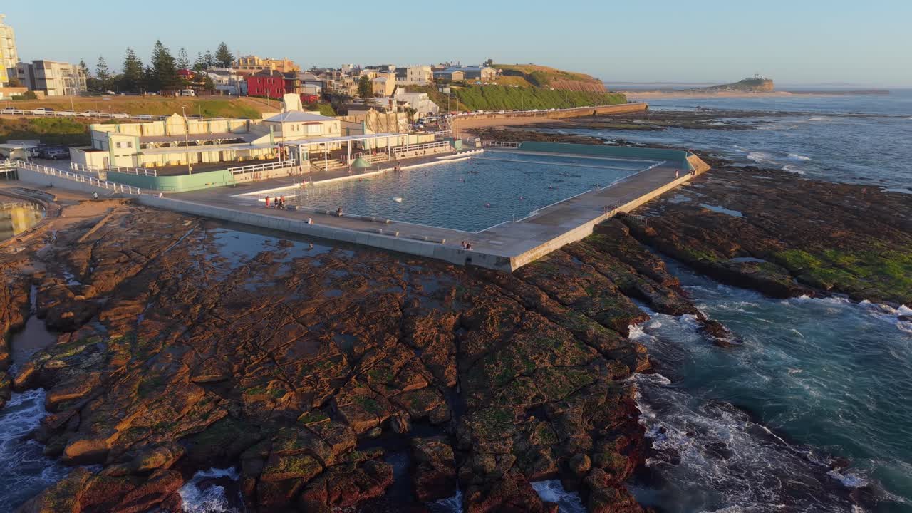 Drone establishing orbit of Newcastle Ocean Baths and rocky shoreline with ocean waves crashing